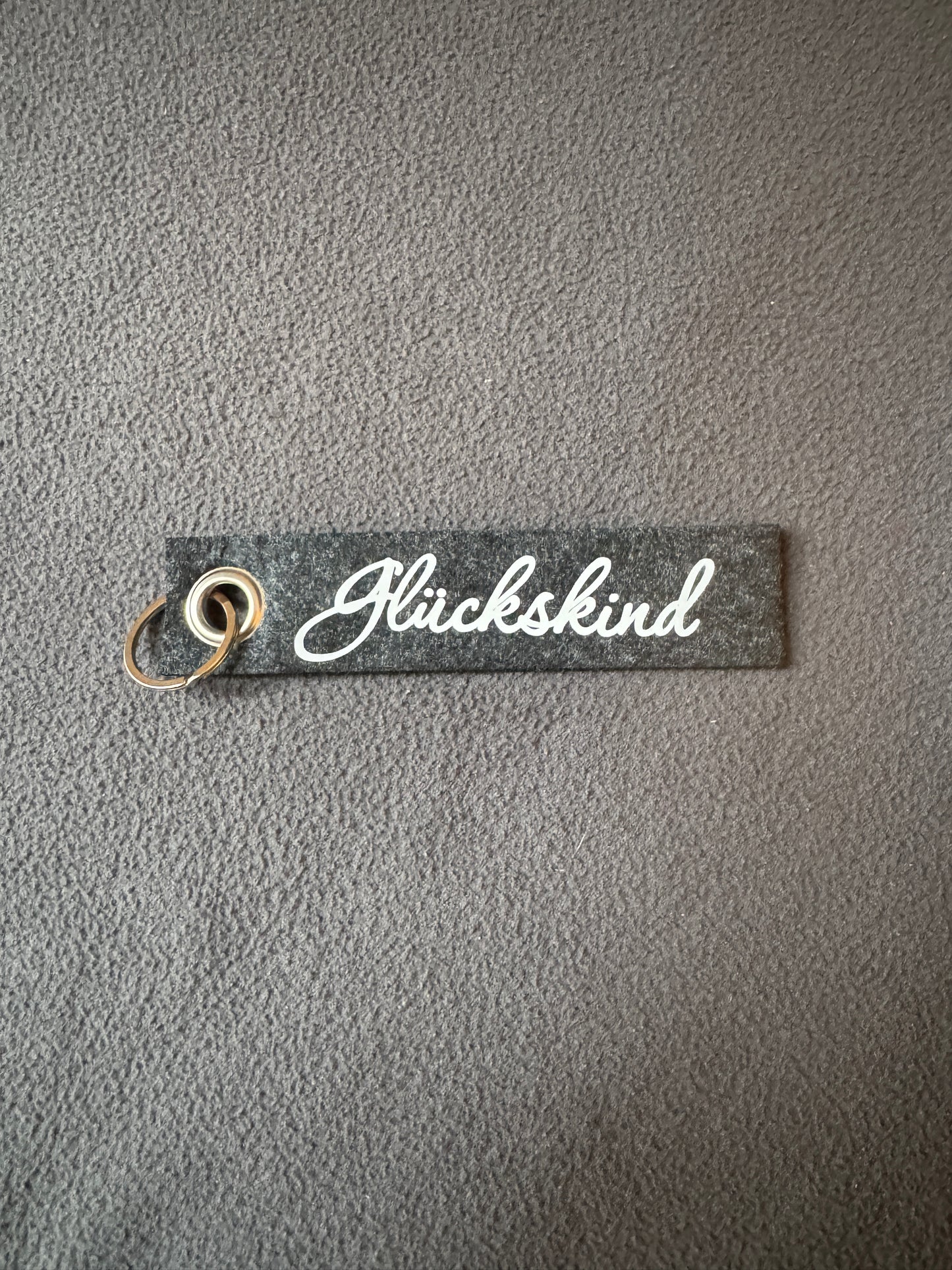 Filzanhänger in dunkelgrau mit weißem Schriftzug „Glückskind“.