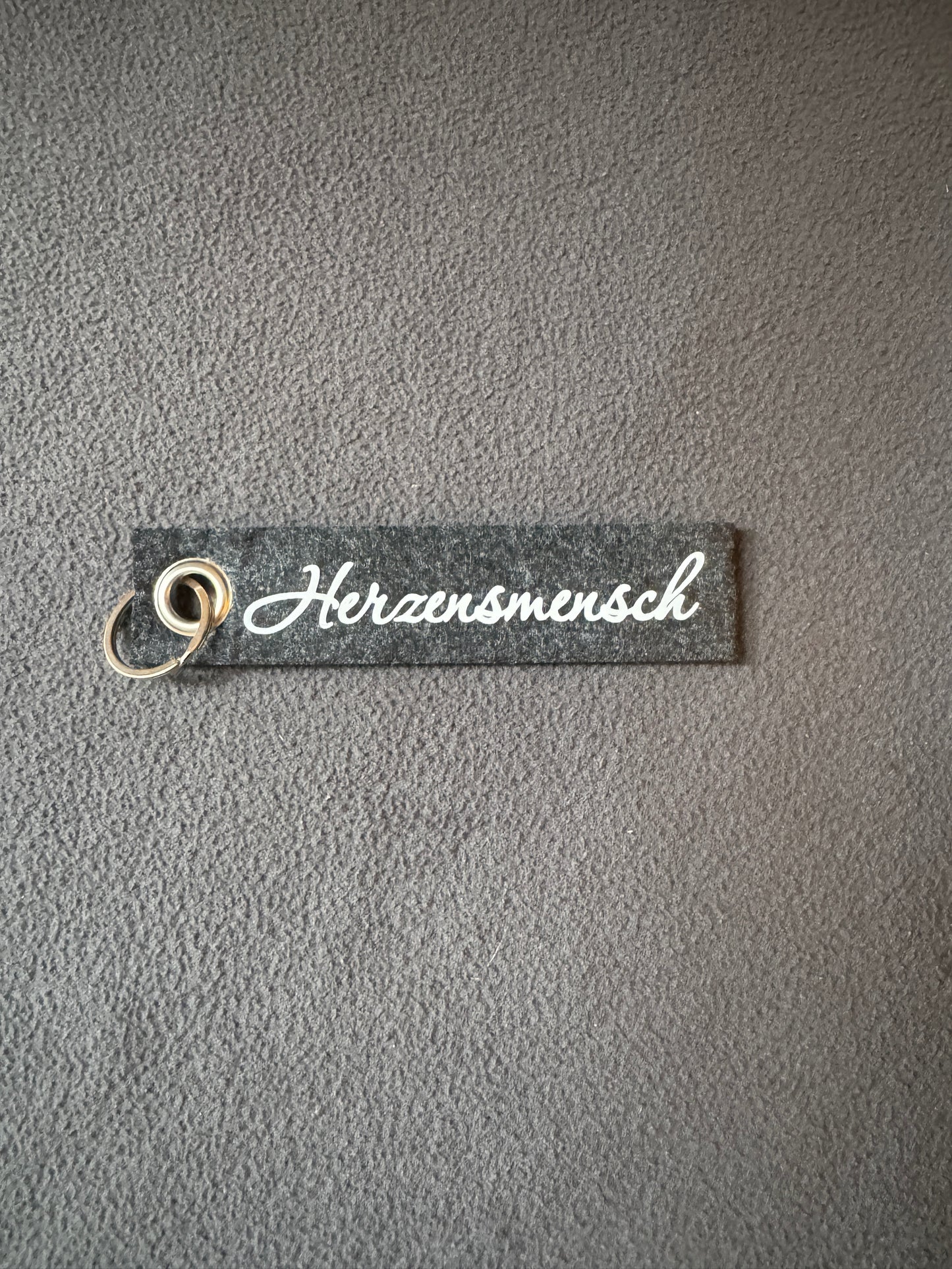 Grauer Filz-Schlüsselanhänger mit dem Schriftzug „Herzensmensch“.
