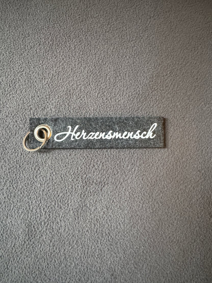 Grauer Filz-Schlüsselanhänger mit dem Schriftzug „Herzensmensch“.
